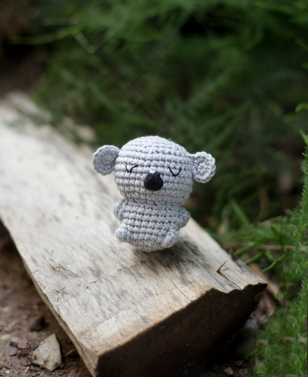 Light grey crochet koala mini with big round ears and a black nose sitting on a piece of driftwood outdoors