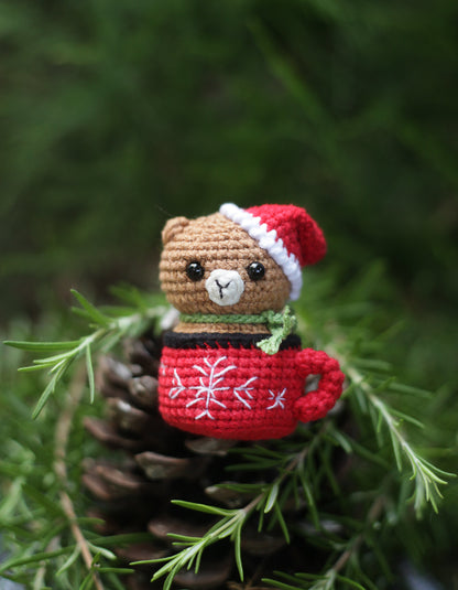 Hand-crocheted brown bear in a Santa hat sitting in a red snowflake mug on a pine cone with greenery