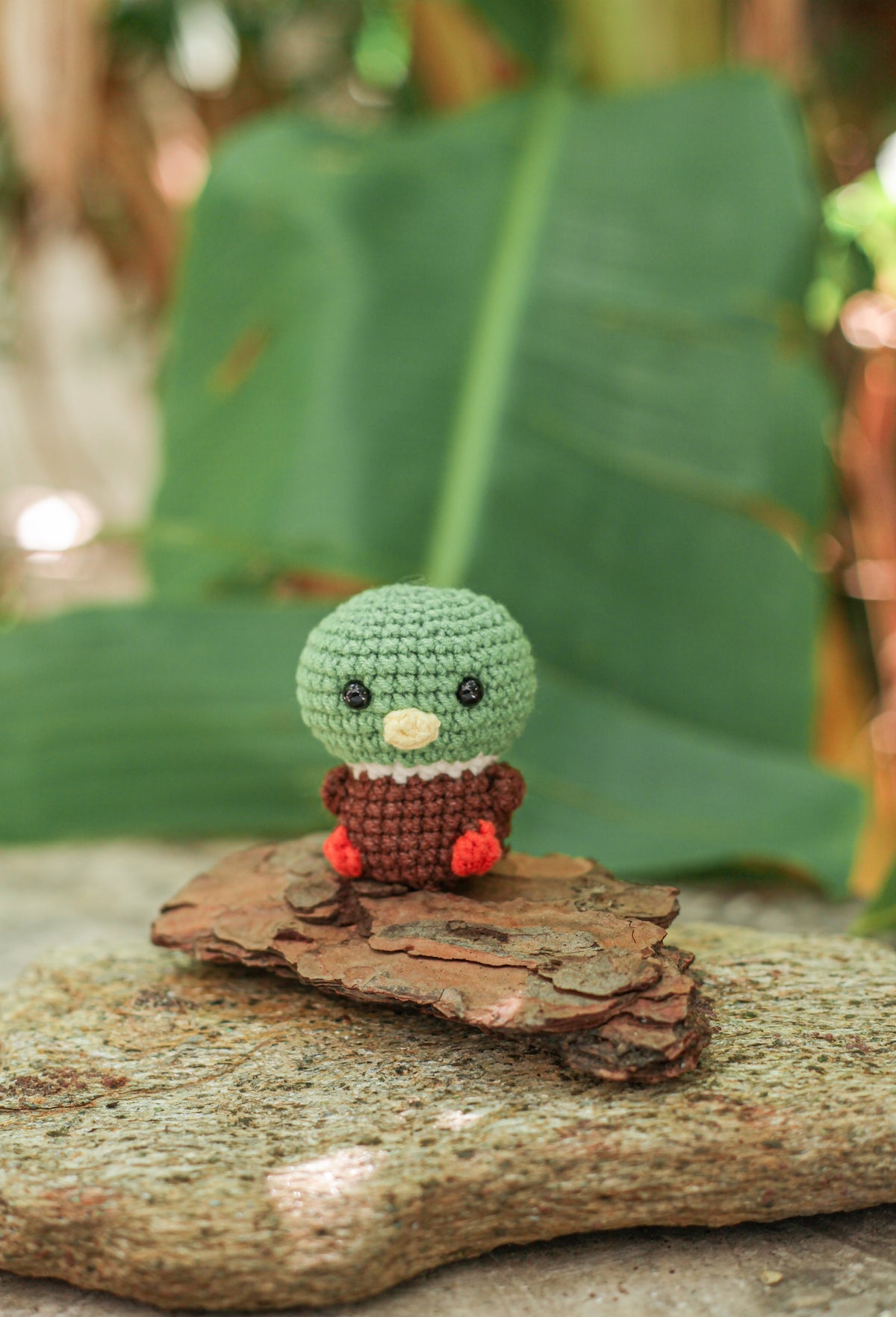 Crochet mallard duck mini with a green head, rust-brown body and red feet sitting on bark in front of a large green leaf