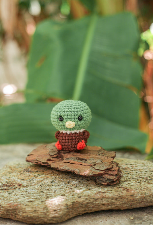 Crochet mallard duck mini with a green head, rust-brown body and red feet sitting on bark in front of a large green leaf