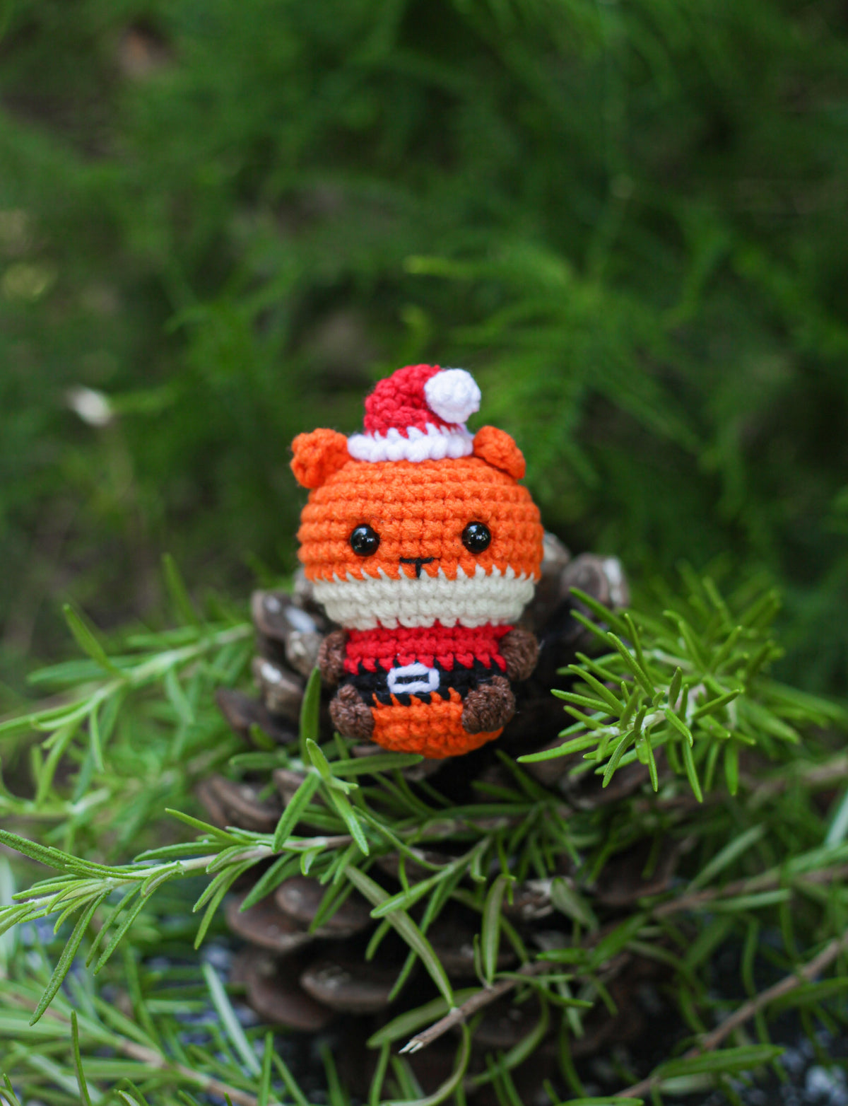 Orange crochet fox mini wearing a Santa hat and red Santa outfit, sitting among pine greenery on a pine cone