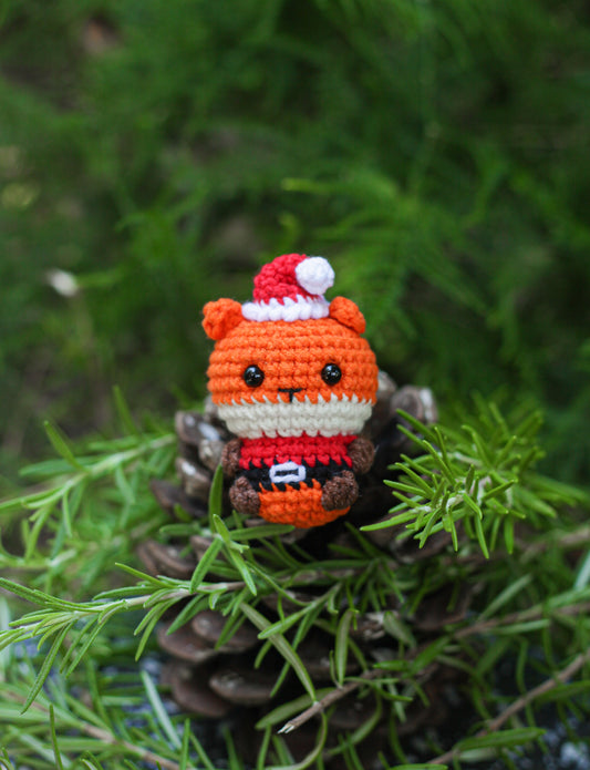 Orange crochet fox mini wearing a Santa hat and red Santa outfit, sitting among pine greenery on a pine cone