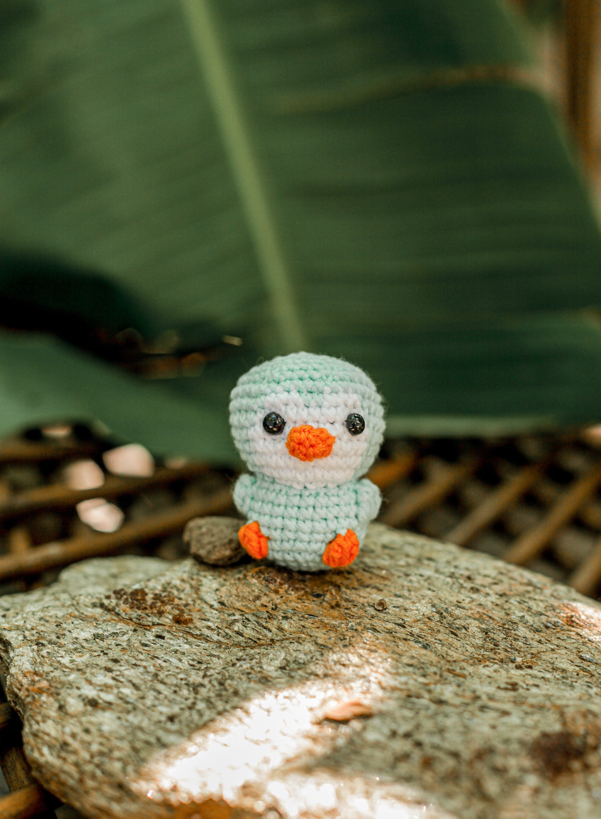 Pale mint and white crochet duck mini with an orange beak and feet standing on a rock in front of a large green leaf