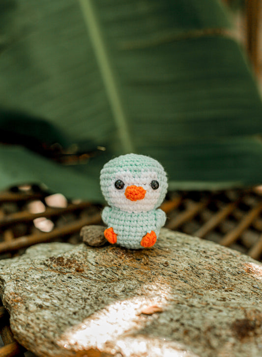 Pale mint and white crochet duck mini with an orange beak and feet standing on a rock in front of a large green leaf