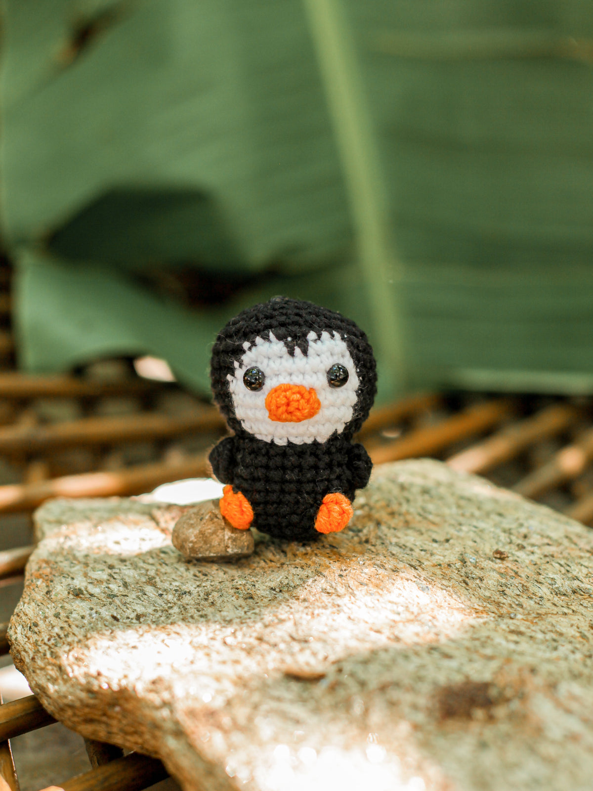 Black crochet penguin mini with a white belly and orange beak and feet sitting on a sunlit rock outdoors