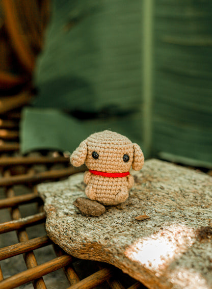 Small sandy-brown crochet bear mini with drooping ears and a red collar, sitting on a rock in front of a large green leaf