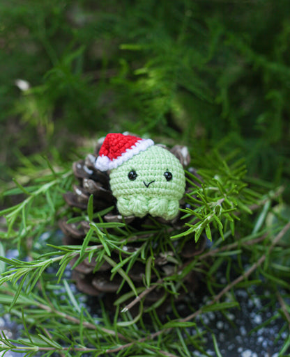 Light green crochet frog wearing a red Santa hat, sitting on a pinecone among evergreen branches