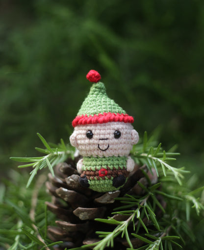 Crochet Christmas elf mini with a green pointed hat and red pom-pom, sitting on a pine cone among greenery