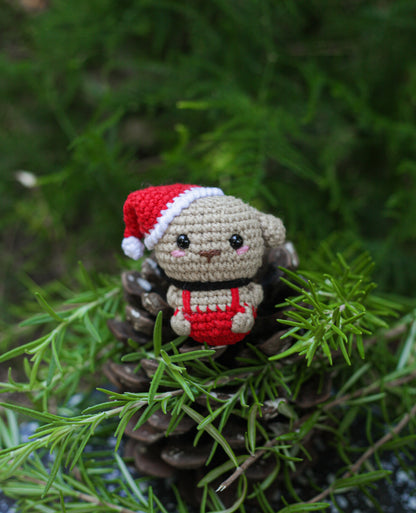 Beige crochet dog mini wearing a Santa hat and red outfit, sitting on a pine cone among pine greenery