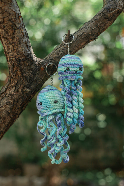Two blue crochet jellyfish keychains with long curly tentacles hanging from a tree branch outdoors