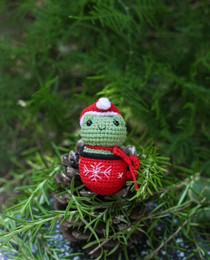 Green crochet frog mini wearing a Santa hat and red scarf with a red snowflake bauble, nestled in pine greenery