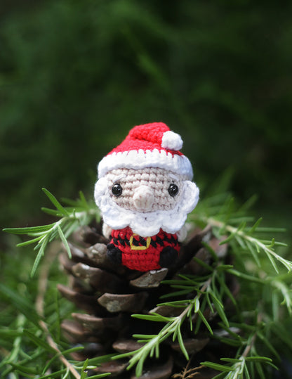 Crochet Santa Claus mini with a red hat and suit and white beard sitting among pine cones and greenery