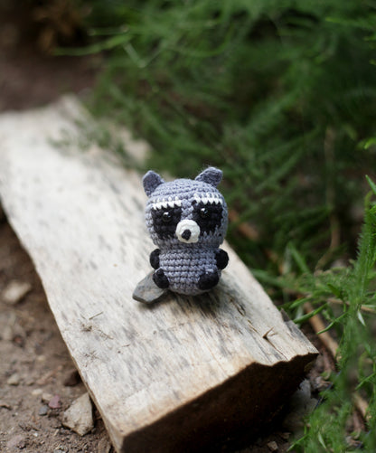 Grey crochet raccoon mini sitting on a fallen log outdoors among green foliage