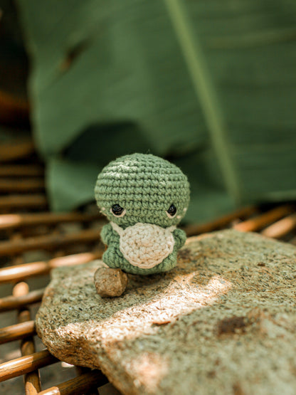 Green crochet turtle mini with a cream belly sitting on a stone with a woven basket background