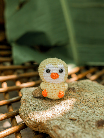 Pale yellow crochet duck keychain with an orange beak and feet, perched on a rock outdoors