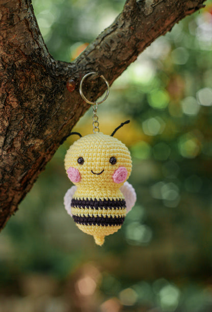 Yellow and black striped crochet bee keychain with pink cheeks hanging from a tree branch
