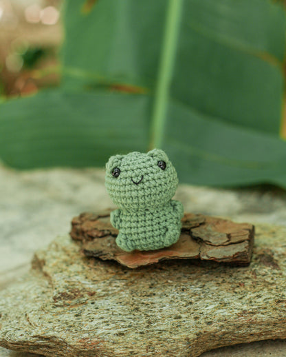 Light green crochet frog mini sitting on bark with a large green leaf behind it