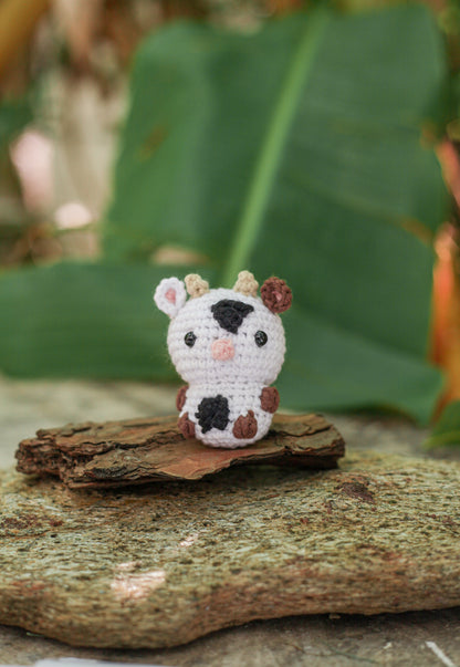 White crochet cow mini with black patches, tiny horns and a pink nose, sitting on bark outdoors