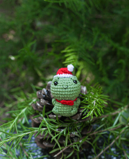 Green crochet frog mini wearing a red Santa hat, nestled among pine branches and pinecones