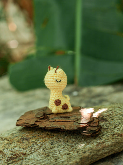 Small pale-yellow crochet giraffe mini with brown spots sitting on a piece of bark outdoors with green leaves behind