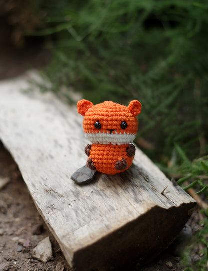 Orange crochet fox mini with a cream muzzle and brown paws, sitting on a piece of wood outdoors