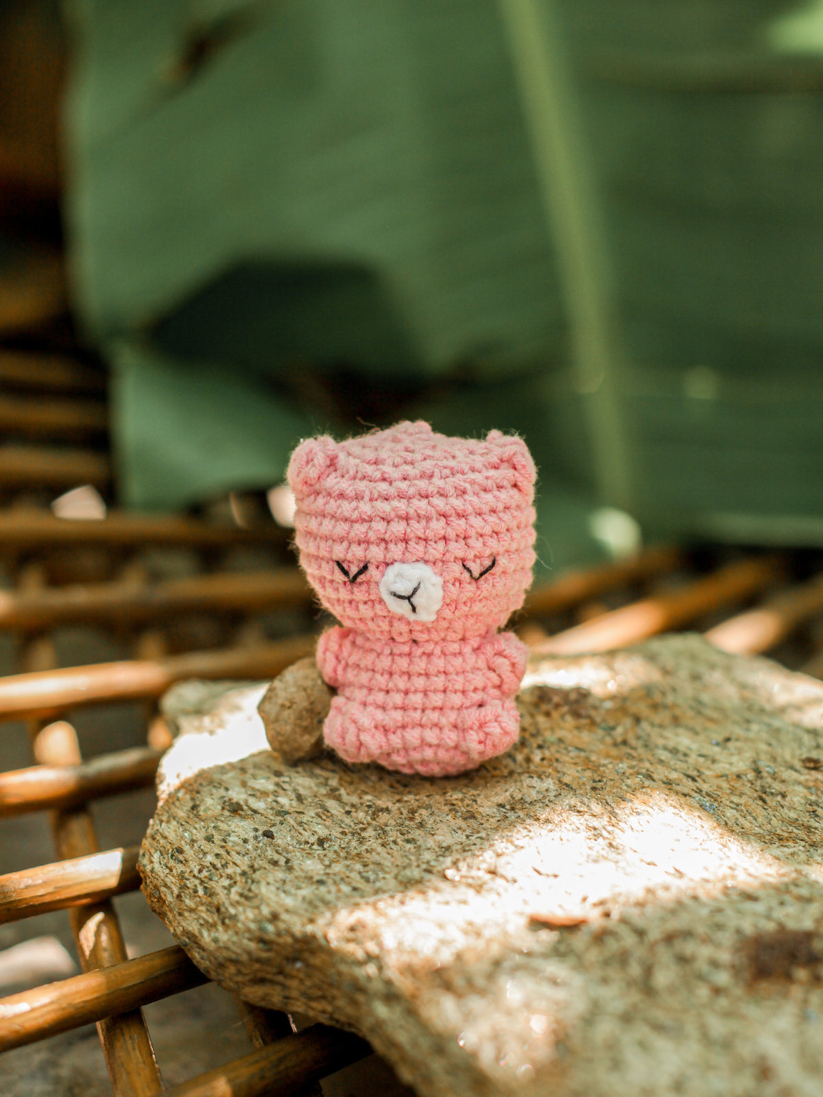 Pink crochet bear mini with embroidered closed eyes sitting on a sunlit rock with large green leaves behind it