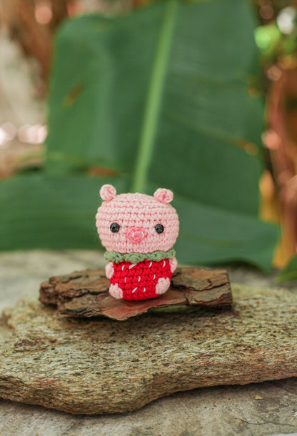 Pink crochet pig mini in a red strawberry-print outfit, perched on bark with green leaves behind