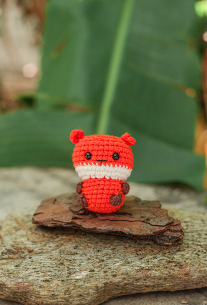 Bright red-orange crochet fox mini with a white muzzle and brown paws, standing on bark in front of green leaves