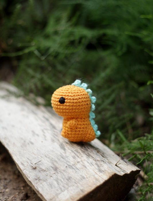 Orange crochet dinosaur mini with pale blue spikes along its back standing on a piece of driftwood outdoors