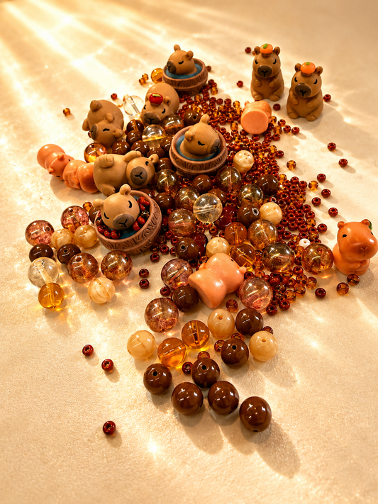 Caspian bead box mix spread on cream fabric — brown and amber beads with small capybara charms.