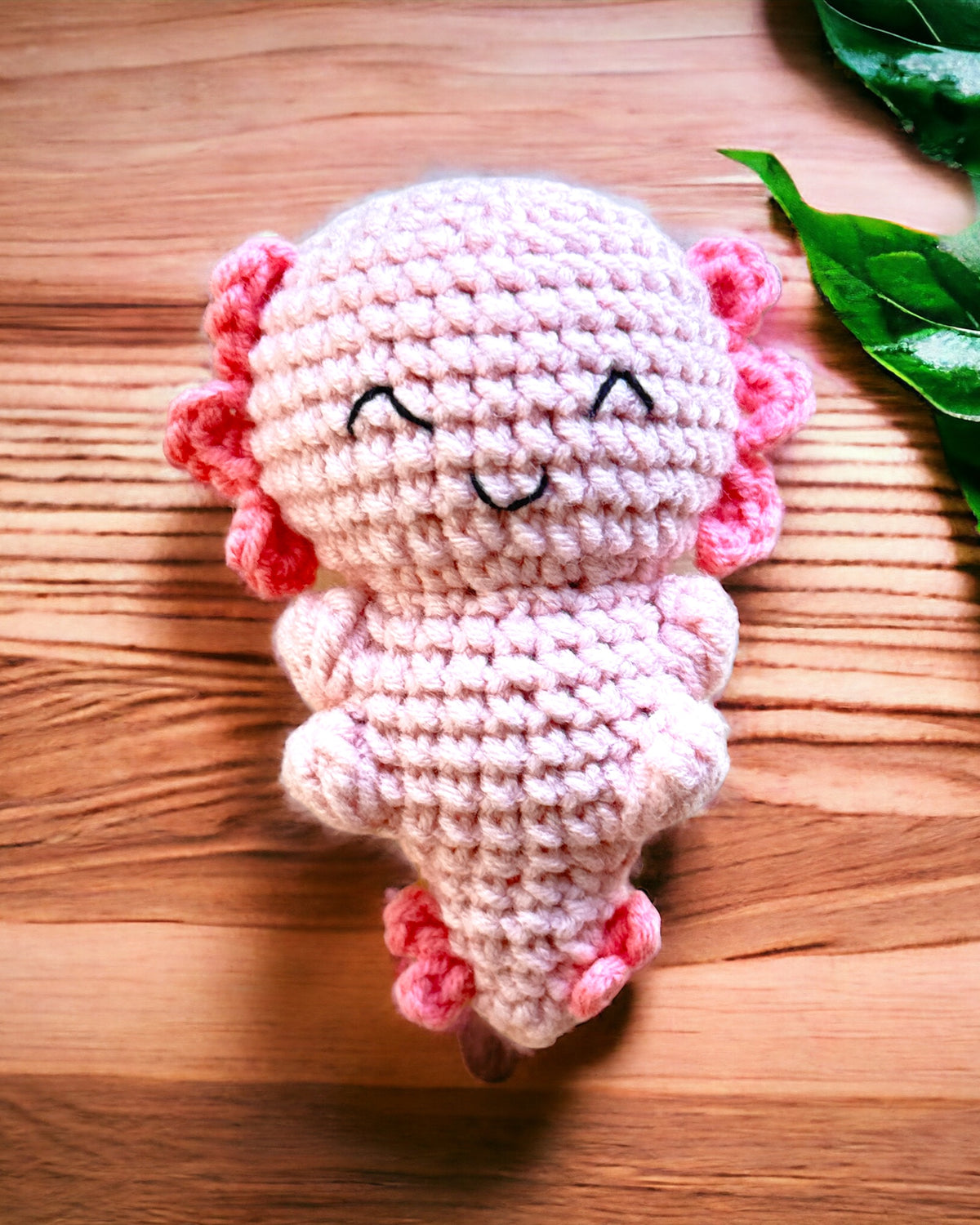 Pink crochet axolotl mini with darker pink feathery gills, lying on a wooden table with a stitched smile